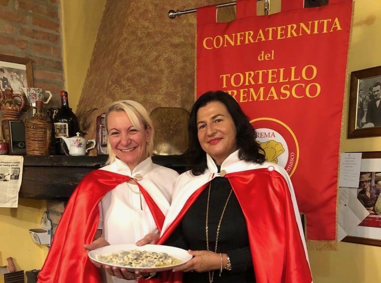 Piatto di tortello cremasco De.Co. con due membri della Confraternita del Tortello Cremasco in mantella rossa e bianca e stemma ufficiale sullo sfondo